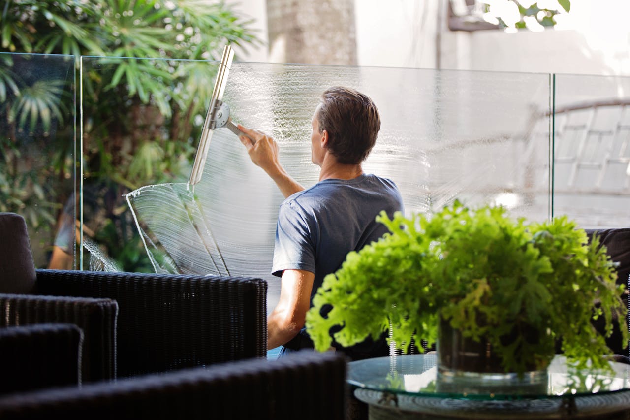 Services A man cleaning a glass barrier outdoors with a squeegee, surrounded by greenery.